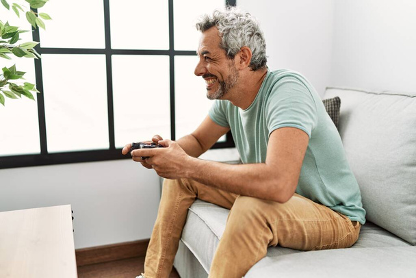 Middle age grey-haired man playing video game sitting on sofa at home - Photo, image