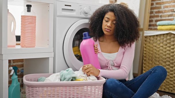 Afričanky americká žena mytí oblečení drží čisticí prostředek láhev se smutným výrazem v prádelně - Fotografie, Obrázek