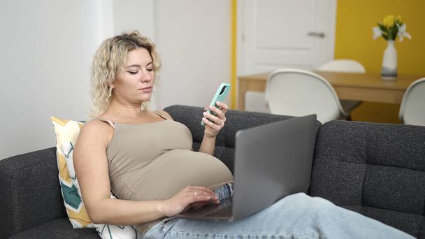 Nuori raskaana oleva nainen, joka käyttää älypuhelinta ja kannettavaa tietokonetta sohvalla kotona - Valokuva, kuva