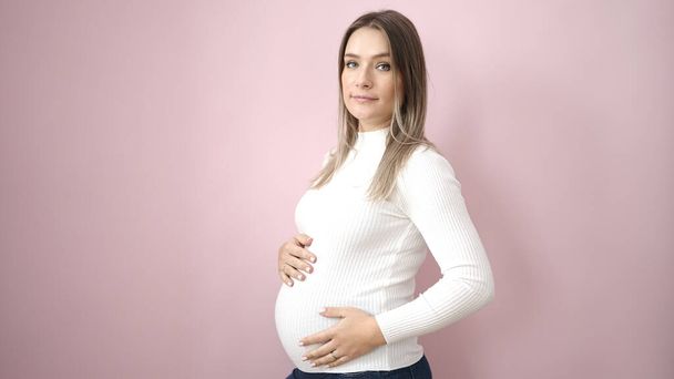 Mujer embarazada joven tocando el vientre con expresión relajada sobre fondo rosa aislado - Foto, imagen