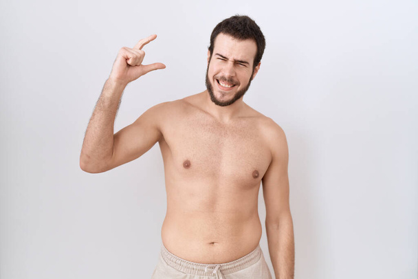 Jonge Spaanse man staat shirtloos over witte achtergrond glimlachend en zelfverzekerd gebaar met de hand doen klein formaat teken met vingers kijken en de camera. meetconcept.  - Foto, afbeelding