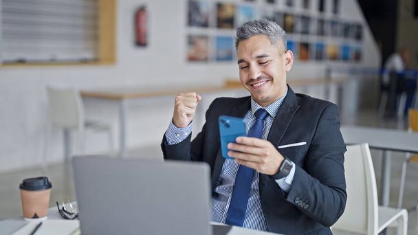 Giovane uomo ispanico imprenditore in cerca di smartphone con gesto vincitore in ufficio - Foto, immagini