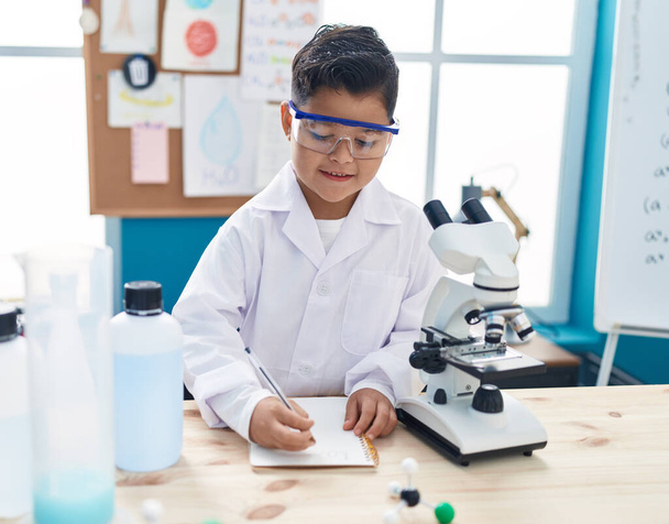 Schattige Spaanse jongen student met behulp van microscoop het schrijven van notities in laboratorium klaslokaal - Foto, afbeelding