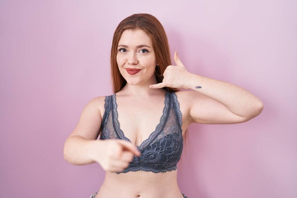 Redhead woman wearing lingerie over pink background smiling doing talking on the telephone gesture and pointing to you. call me.  - Photo, image