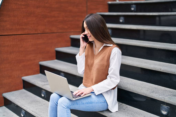 Молода красива іспаномовна жінка розмовляє на смартфоні за допомогою ноутбука, що сидить на сходах на вулиці
 - Фото, зображення