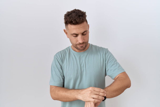Hispánec s plnovousem stojící nad bílým pozadím kontrolující čas na hodinkách na zápěstí, uvolněný a sebejistý  - Fotografie, Obrázek
