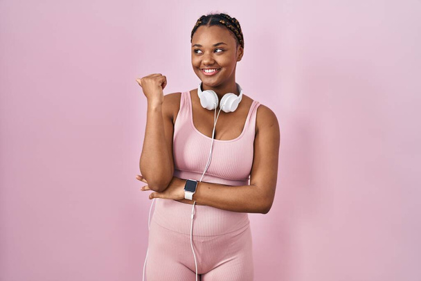 Mujer afroamericana con trenzas con ropa deportiva y auriculares sonriendo con la cara feliz mirando y apuntando hacia un lado con el pulgar hacia arriba.  - Foto, Imagen