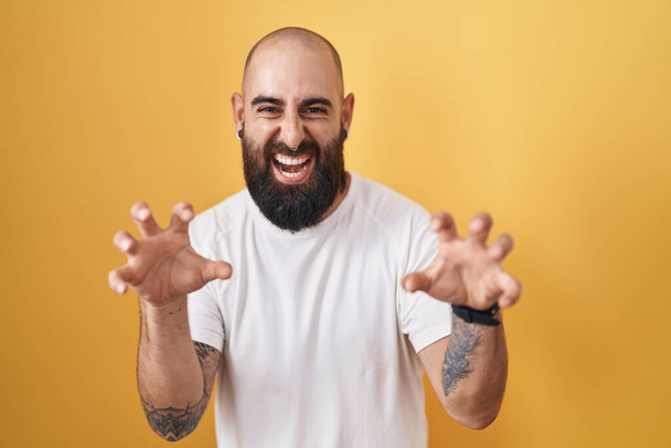 Mladý hispánec s vousy a tetování stojící nad žlutým pozadím s legračním úsměvem dělá drápy gesto jako kočka, agresivní a sexy výraz  - Fotografie, Obrázek