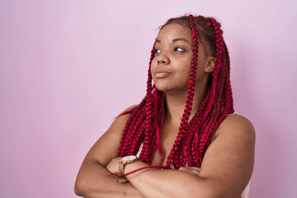 Αφροαμερικανή γυναίκα με πλεγμένα μαλλιά στέκεται πάνω από ροζ φόντο κοιτάζοντας στο πλάι με τα χέρια σταυρωμένα πεπεισμένη και με αυτοπεποίθηση  - Φωτογραφία, εικόνα