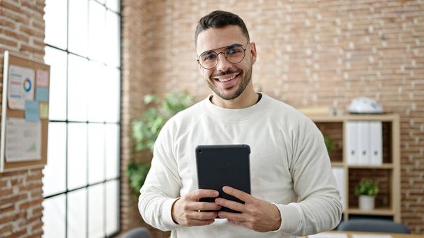Молодий іспаномовний чоловік бізнесмен посміхається впевнено використовуючи тачпад в офісі
 - Фото, зображення
