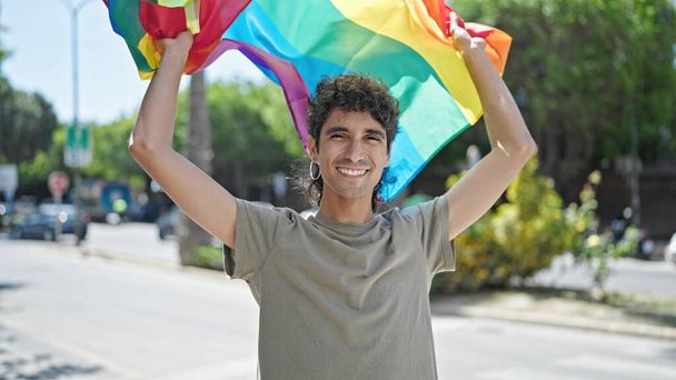 Jonge Spaanse man glimlacht vol vertrouwen met regenboogvlag op straat - Foto, afbeelding