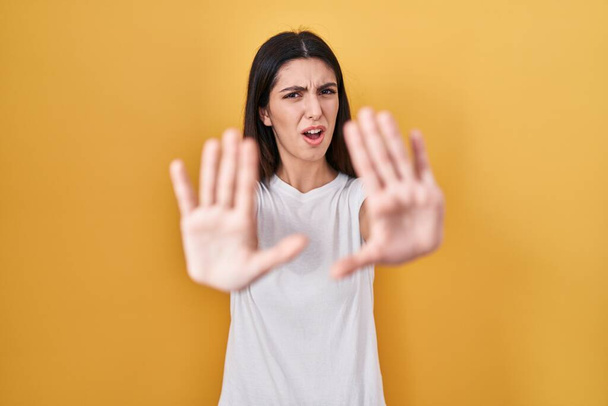 Jovem mulher bonita de pé sobre fundo amarelo fazendo parar gesto com as mãos palmas das mãos, expressão irritada e frustração  - Foto, Imagem