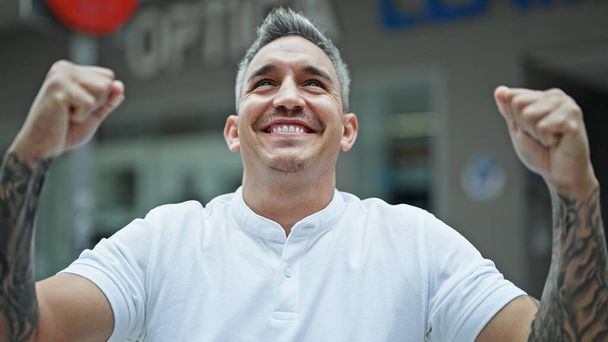Young hispanic man smiling confident looking to the sky with winner gesture at street - Photo, Image