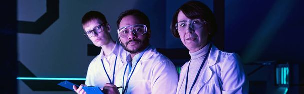 banner, Drie Wetenschappers Betrokken bij Headshot Analyse Binnen Neon-Lit Science Center. - Foto, afbeelding