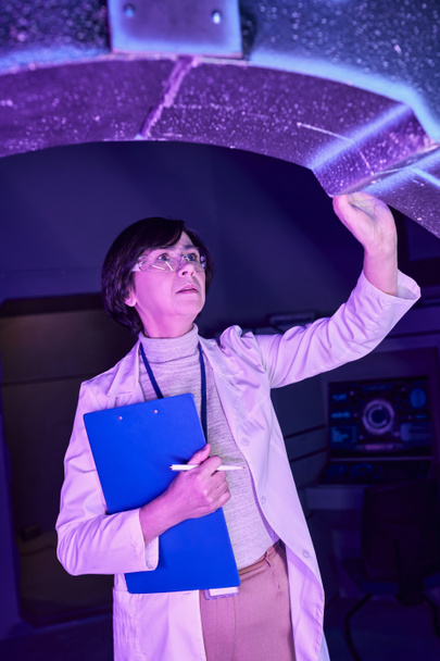 centro de ciencia futurista, científica femenina con portapapeles examinando dispositivo innovador - Foto, Imagen