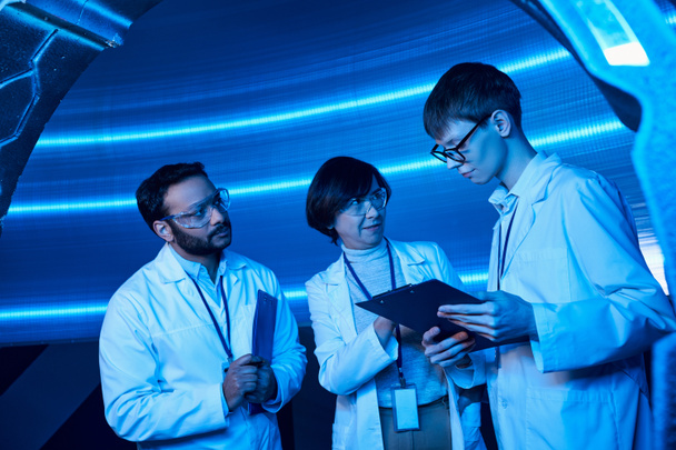 científicos multiétnicos mirando a un joven interno con portapapeles en un centro de ciencia futurista iluminado por neón - Foto, Imagen