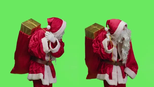 Ojciec Boże Narodzenie ucieleśnienie niesie torbę z pudełkami prezentami w studio, zachowując się jak Święty Mikołaj z prezentami przed zielonym ekranem. Młody dorosły z wielkim workiem zabawek, święto. - Materiał filmowy, wideo