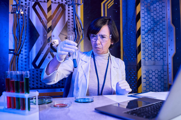 femme scientifique en lunettes regardant le tube à essai avec du liquide près d'un ordinateur portable dans un laboratoire futuriste - Photo, image