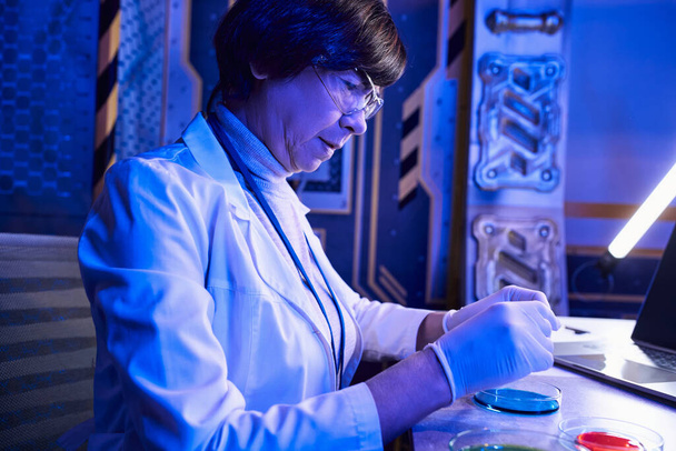 研究室のペトリ皿でエイリアンの生命サンプルを扱うゴーグルの中年の女性科学者 - 写真・画像