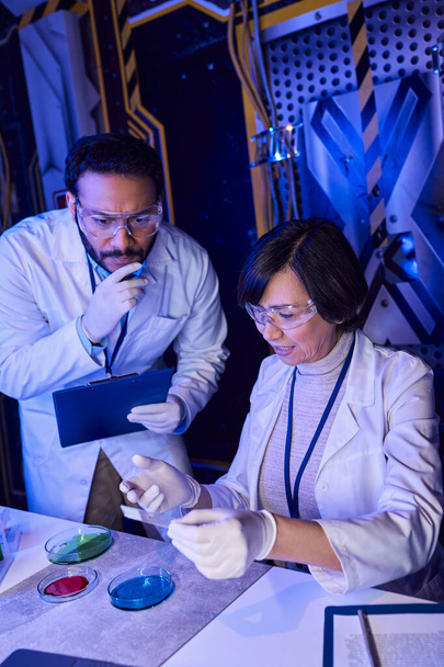 Indiaas wetenschapper met klembord kijken naar laboratoriumglas in de buurt van collega en petrischaaltjes - Foto, afbeelding