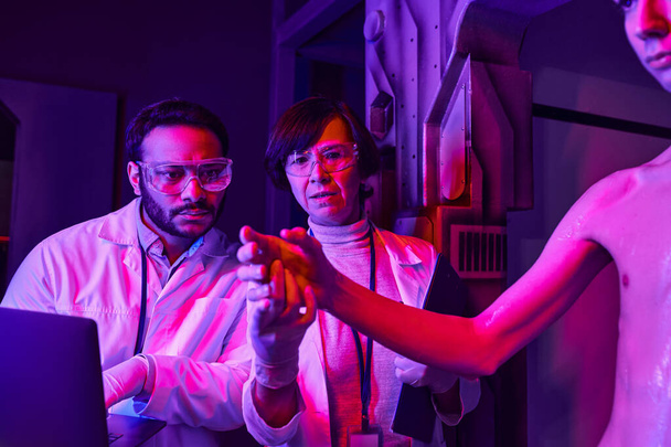 индийский ученый с ноутбуком рядом с коллегой держит за руку космического инопланетянина в инновационной лаборатории - Фото, изображение