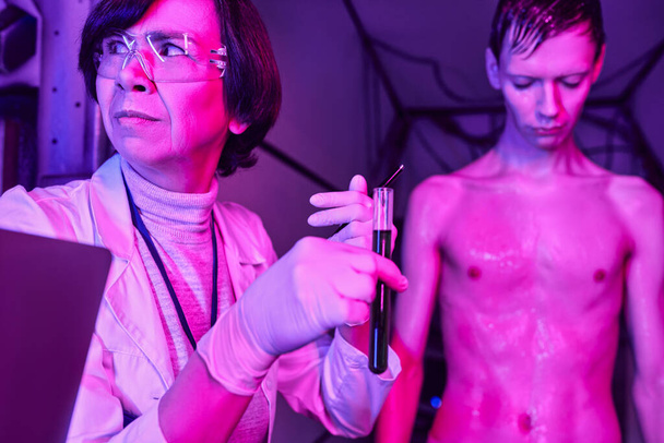 γυναίκα επιστήμονας σε γυαλιά κρατώντας δοκιμαστικό σωλήνα με δείγμα αίματος κοντά στο ανθρωποειδές αλλοδαπός σε καινοτόμο εργαστήριο - Φωτογραφία, εικόνα