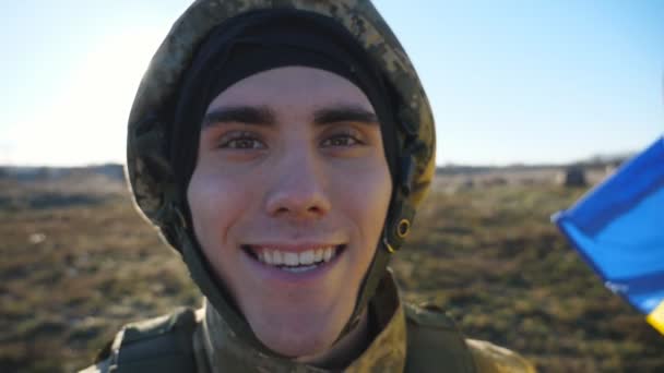 Portrait of happy male ukrainian army soldier in helmet looking into camera outdoor. Positive gaze of young military man with blue-yellow flag at countryside. Invasion of territory Ukraine. End of war - Footage, Video