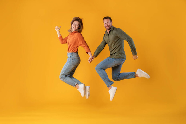 Aufgeregte junge europäische Ehepartner rennen, springen, frieren in der Luft und lächeln in die Kamera, isoliert auf gelbem Hintergrund, in voller Länge. Gemeinsamer Spaß, Angebot und Werbung, Verkauf - Foto, Bild