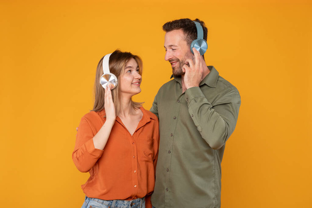 Enjoy music together. Cheerful young couple using wireless headphones, listening favourite songs and looking at each other, isolated on orange background - Фото, изображение