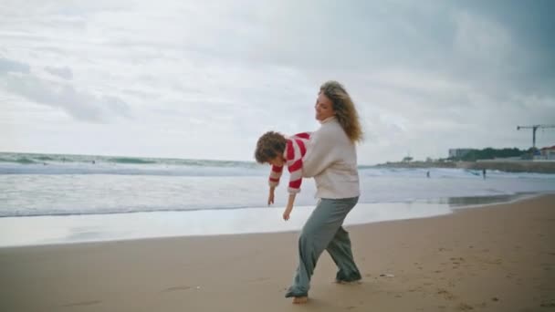 Vidám anya forog gyerek őszi tengerparton. Mosolygó bébiszitter, aki göndör fiút tart a kezében. Gyönyörű fiatal anya játékos fia szórakozás együtt a tengerparton. Szerető családi kapcsolatok - Felvétel, videó