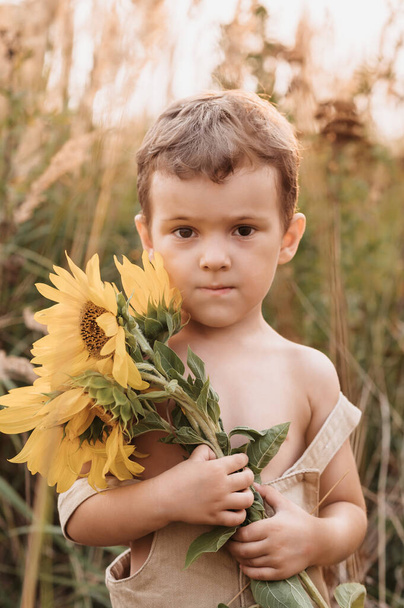 Un lindo niño riendo en un campo de girasoles, sosteniendo un enorme ramo de flores en sus manos en una soleada noche de verano. La hora dorada - Foto, imagen