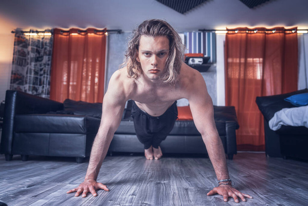 Νεαρός μοντέλο αθλητικός άνθρωπος άσκηση push up στο πάτωμα του εσωτερικού στούντιο σπίτι του. Μυώδης καυκάσιος αγόρι προπονεί push-ups αθλητική ρουτίνα κοιτάζοντας την κάμερα δείχνει τα χέρια και τους κοιλιακούς από το όμορφο σώμα του - Φωτογραφία, εικόνα