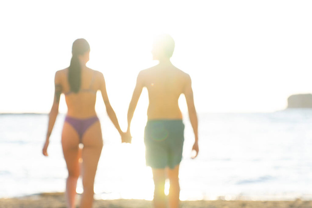 Jóképű férfi fürdőruhát visel, gyönyörű nővel sétál a parton egy napsütéses napon, kézen fogva, ünnepek alatt. Fiatal felnőttek kaukázusi pár utaznak együtt egy romantikus nyaralás pihentető szabadban - Fotó, kép