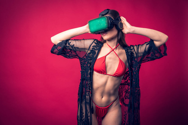 Studio photo shoot of beautiful brown hair model posing wearing red swimwear and vr glasses with red background behind. Sexy woman wearing futuristic augmented reality goggles - Foto, Bild