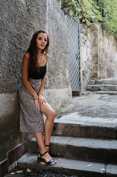 Une jeune brune pose contre un mur de pierre à côté des escaliers. Une belle et mince silhouette. Une fille en robe - Photo, image