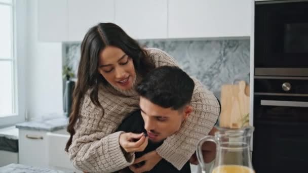 Positive lovers having fun at morning kitchen close up. Happy woman feeding man with berries at home. Latina spouses relaxing at modern interior. Handsome brunette guy piggyback wife at cozy house - Footage, Video