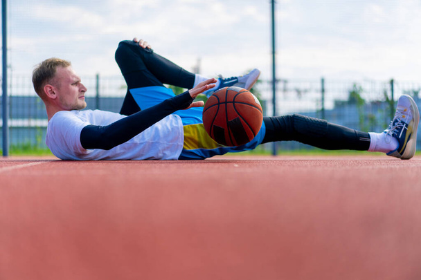Uzun boylu basketbol oyuncusu dışarıda bir basketbol sahasında yatıyor ve antrenman başlamadan önce kaslarını esnetiyor ve topu dolduruyor. - Fotoğraf, Görsel