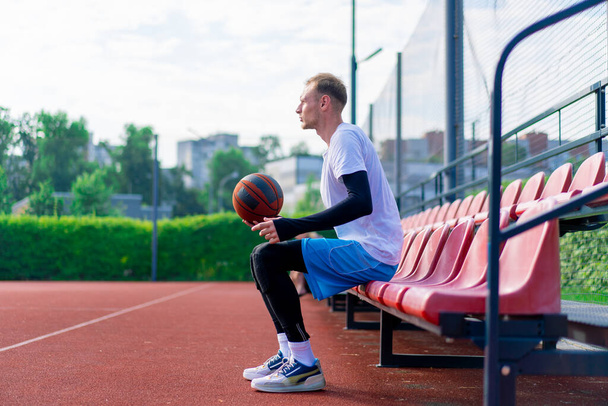 Tall guy basketball player sits in the bleachers of an outdoor basketball court with ball in hand before practice starts - Photo, Image