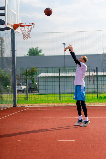высокий парень баскетболист бросает мяч в баскетбольное кольцо на баскетбольной площадке в парке во время тренировки - Фото, изображение