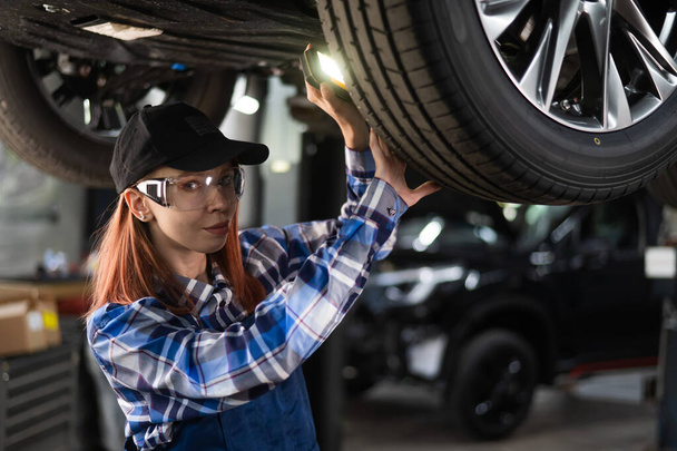 A female mechanic inspects a lifted car. A girl at a mans work - Zdjęcie, obraz