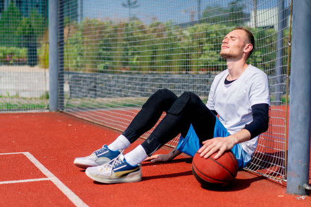 Високий хлопець баскетболіст з м'ячем у руці сидить спираючись на сітку баскетбольного майданчика під час практики в парку - Фото, зображення