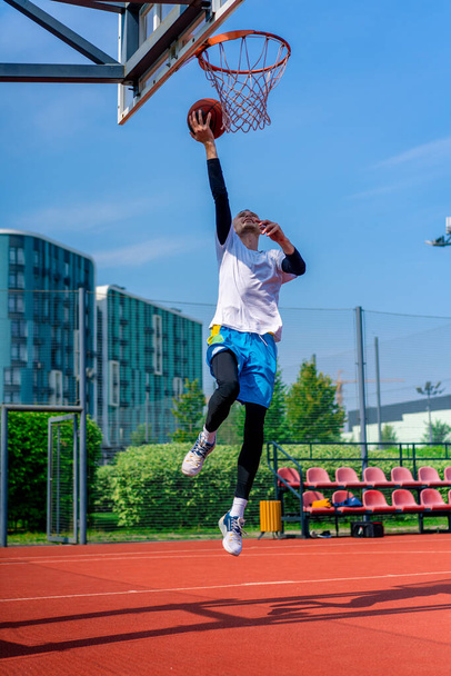 Высокий парень баскетболист прыгает на обруч с мячом в руке, чтобы забить впечатляющий бросок во время тренировки на баскетбольной площадке в парке - Фото, изображение