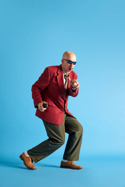 Obraz v celé délce. Plešatý muž s knírem oblečený v obchodním retro obleku plíží přes modré pozadí studia. Pojem emoce, úspěch, retro, retro a reklama - Fotografie, Obrázek