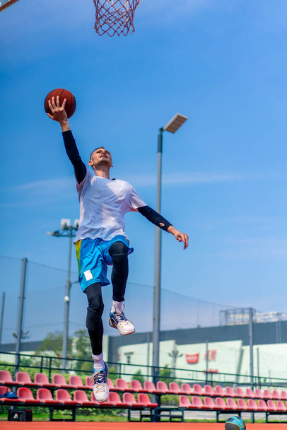 Высокий парень баскетболист прыгает на обруч с мячом в руке, чтобы забить впечатляющий бросок во время тренировки на баскетбольной площадке в парке - Фото, изображение
