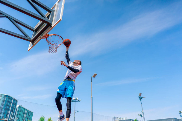 Lange man basketbalspeler springt naar de ring met de bal in zijn hand om een spectaculaire dunk te scoren tijdens de training op het basketbalveld in het park - Foto, afbeelding