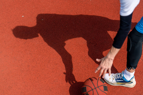 Közelkép a kosárlabdás srác árnyékáról a kosárlabda pálya padlóján, miközben csöpög a labda. - Fotó, kép