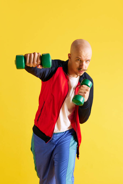 Вертикальная съемка человека, одетого в спортивную одежду и делающего упражнения с ярко-зелеными манекенами, смотрящего на камеру на желтом фоне. Концепция спорта, здорового образа жизни, тренировок и рекламы - Фото, изображение