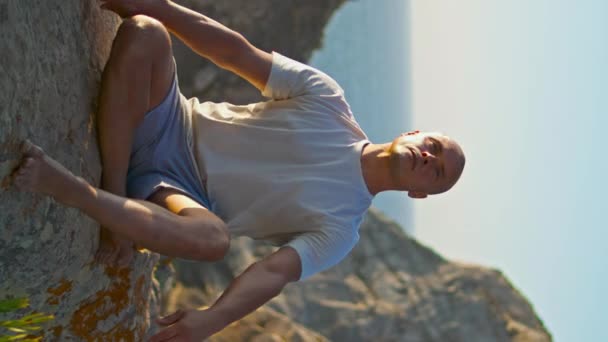 Muscular man practicing asana in morning sunlight. Athlete train at ocean cliff stretching on rock outdoors. Serene strong person relaxing breathing in yoga position vertical view. Fitness recreation. - Footage, Video