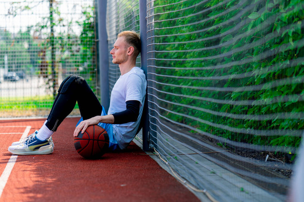 A tall guy basketball player with a ball in his hand sits leaning on the net of a basketball court during practice in park - Photo, Image