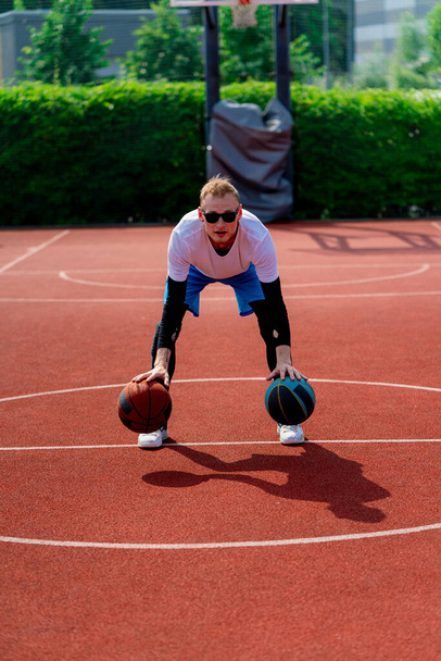 Ένας ψηλός μπασκετμπολίστας με δύο μπάλες επιδεικνύει τις ντρίμπλες ικανότητές του ενώ εξασκείται στο γήπεδο μπάσκετ στο πάρκο. - Φωτογραφία, εικόνα
